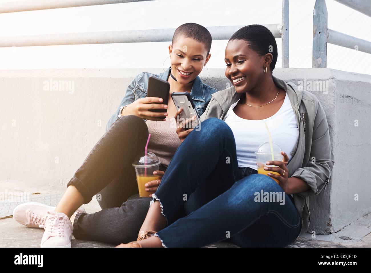 Echte Freunde teilen immer Trends. Aufnahme von zwei jungen Frauen in der Stadt, die lachen und ihre Getränke halten und SMS lesen. Stockfoto