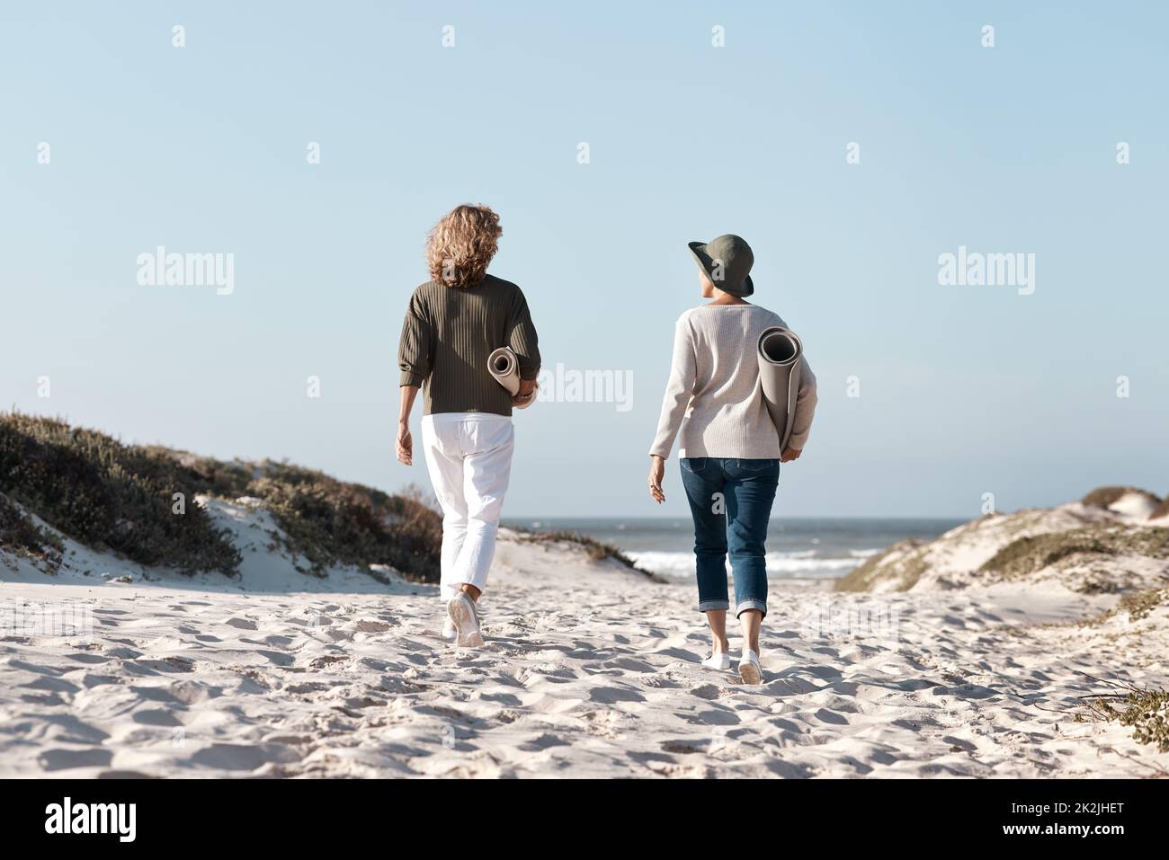 Machen Sie sich auf den Weg, um den perfekten Ort zu finden. Rückansicht von zwei nicht erkennbaren Frauen, die mit ihren Matten am Strand spazieren. Stockfoto