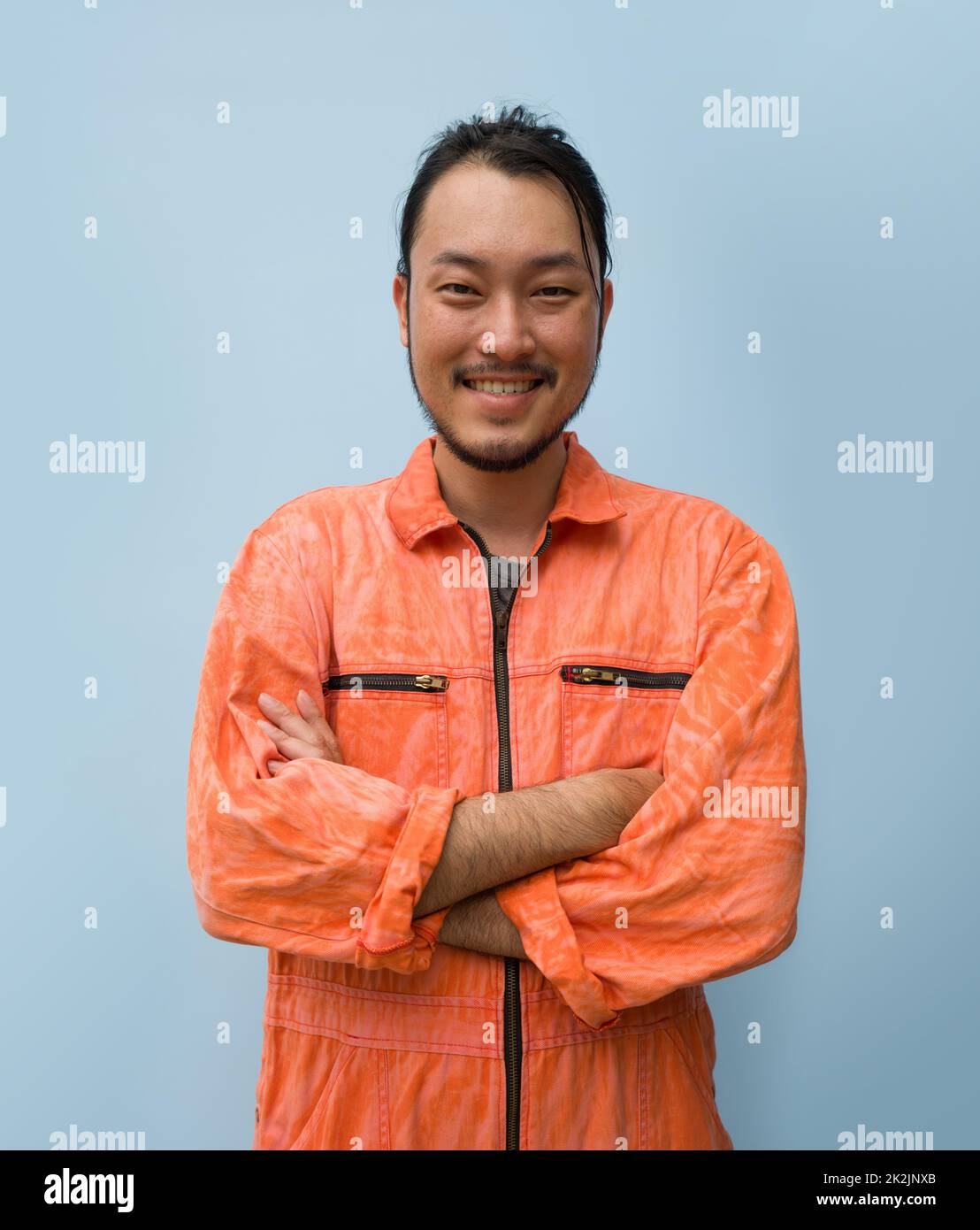 Junger, langhaariger Asiatischer mit Schnurrbart und Bart steht lächelnd mit gekreuzten Armen. Porträt von Industriemenschen auf blauem Hintergrund. Stockfoto