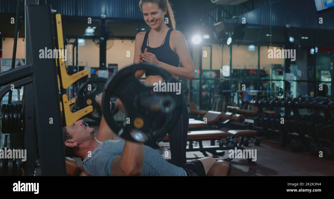 Personal Trainer hilft dem Muskelmann auf der Bank beim Gewichtheben mit der Hakenglocke, die die Muskeln biegt Stockfoto
