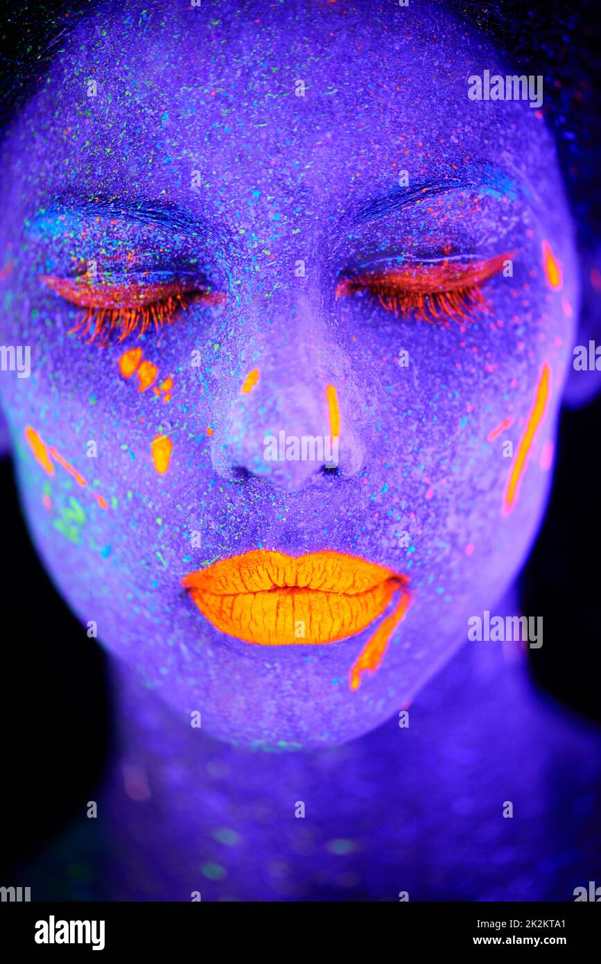 Leuchtende Göttin im Dunkeln. Eine junge Frau posiert mit Neonfarbe auf ihrem Gesicht. Stockfoto
