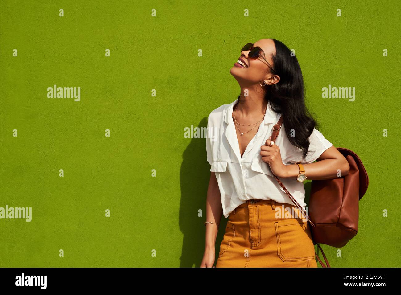 Stilvoll zu bleiben, ist mir wichtig. Gekürzte Aufnahme einer attraktiven jungen Frau mit Sonnenbrille, während sie allein vor einem grünen Hintergrund in der Stadt steht. Stockfoto