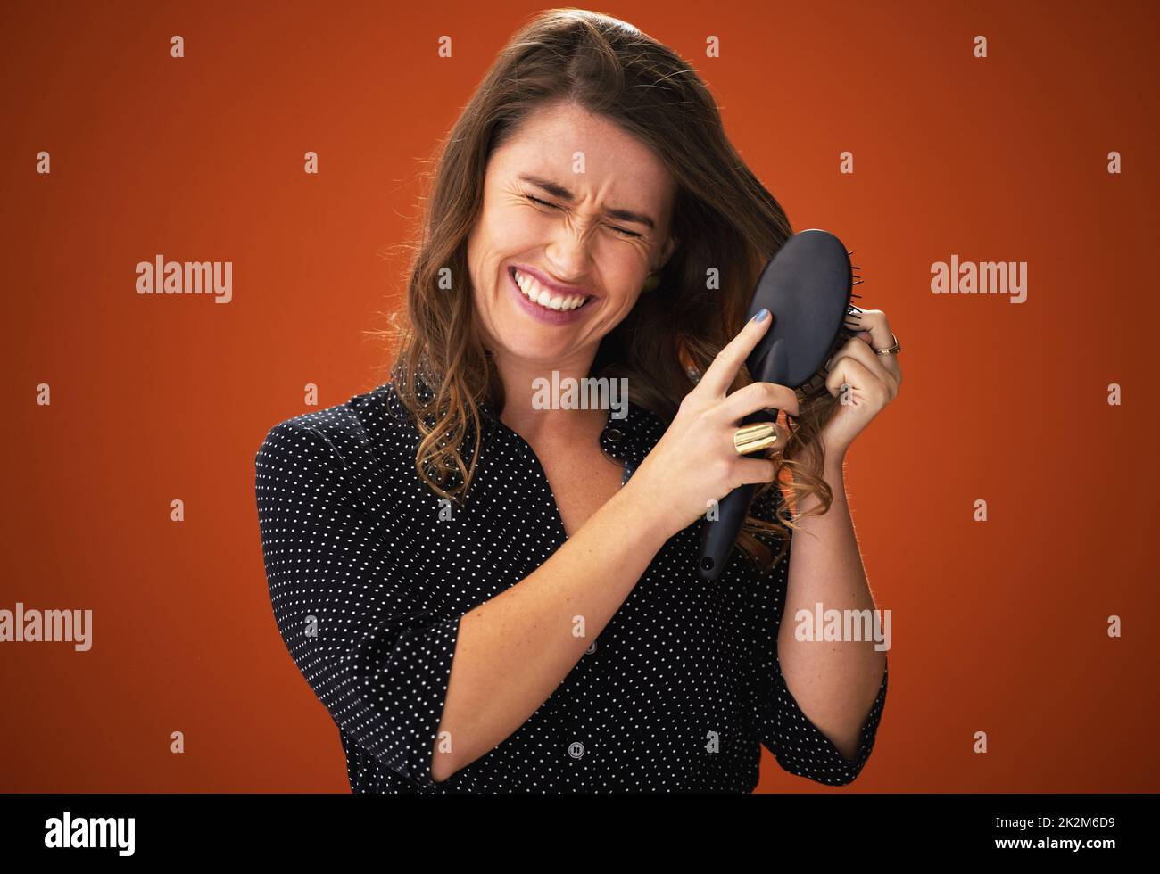 Also... viele... Knoten. Aufnahme einer attraktiven jungen Frau, die im Studio vor einem roten Hintergrund steht und kämpft, um ihr Haar zu putzen. Stockfoto