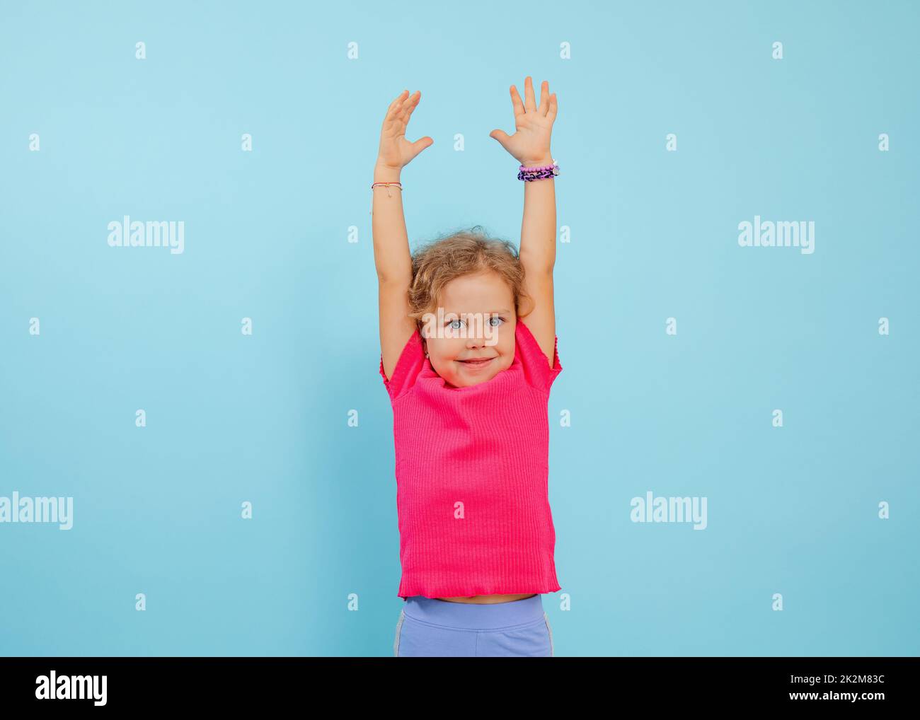 Porträt eines fröhlichen blauäugigen kleinen Mädchens mit lockigem, hellem Haar, das ein rosa T-Shirt trägt und die Hände auf blauem Hintergrund hochhebt. Stockfoto