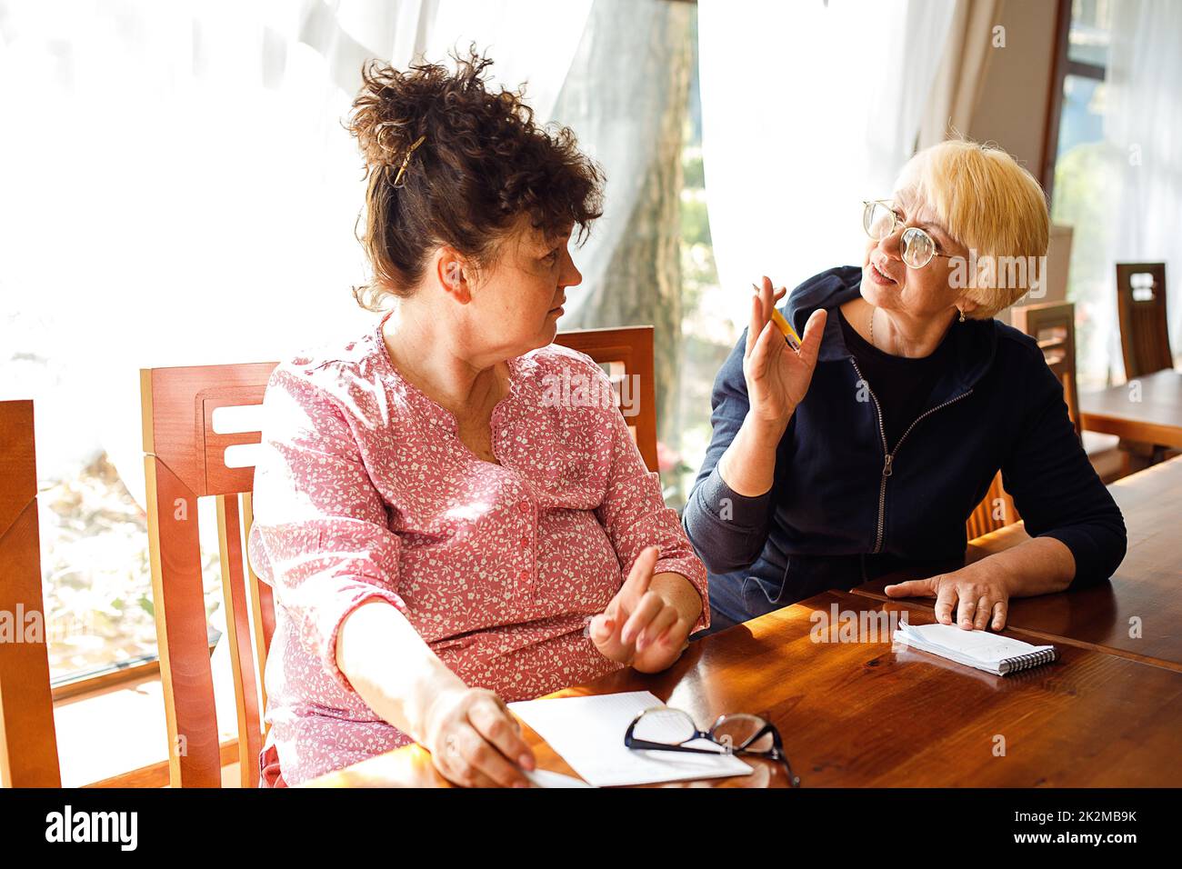 Zwei enthusiastische ältere Frauen tragen legere Kleidung, sitzen am Schreibtisch, diskutieren, machen Notizen in Notizblock im Büro. Stockfoto