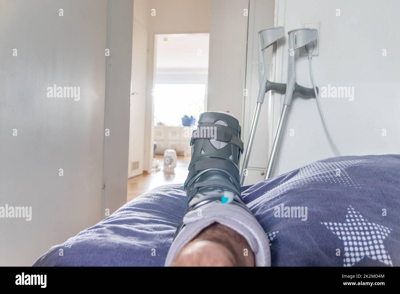 Der europäische Mann nach der Achillessehnenruptur ist wieder zu Hause mit einem speziellen Physiotherapieschuh und Krücken für die Heilung zu Hause mit gesunden Medikamenten Schmerzmittel gegen das schmerzende Bein Stockfoto