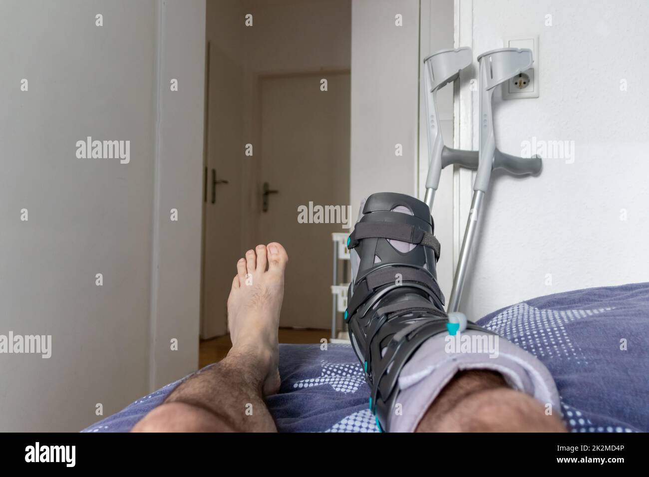 Der europäische Mann nach der Achillessehnenruptur ist wieder zu Hause mit einem speziellen Physiotherapieschuh und Krücken für die Heilung zu Hause mit gesunden Medikamenten Schmerzmittel gegen das schmerzende Bein Stockfoto