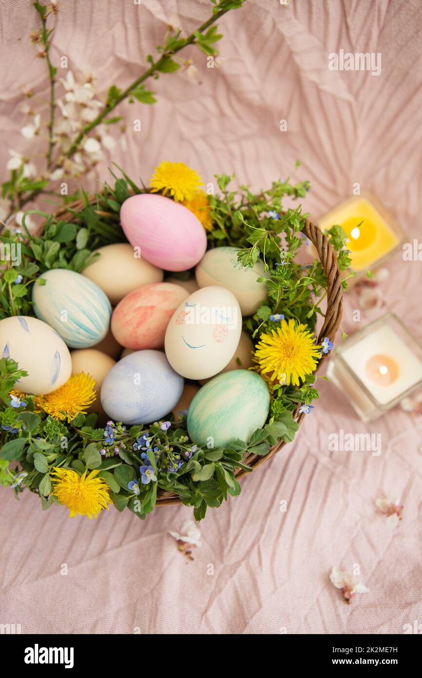 Auf einer pastellfarbenen Bettdecke befindet sich ein Korb mit Gras und leuchtenden Blumen. Der Korb enthält Ostereier, die mit Aquarellen in wunderschönen Pastelltönen bemalt sind. Stockfoto