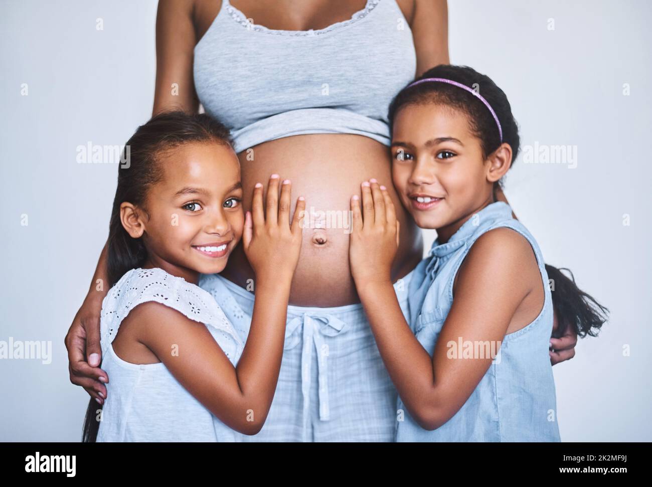 Hören wir mal zu. Porträt von zwei fröhlichen kleinen Mädchen, die tagsüber neben ihrer Schwangeren zu Hause stehen. Stockfoto