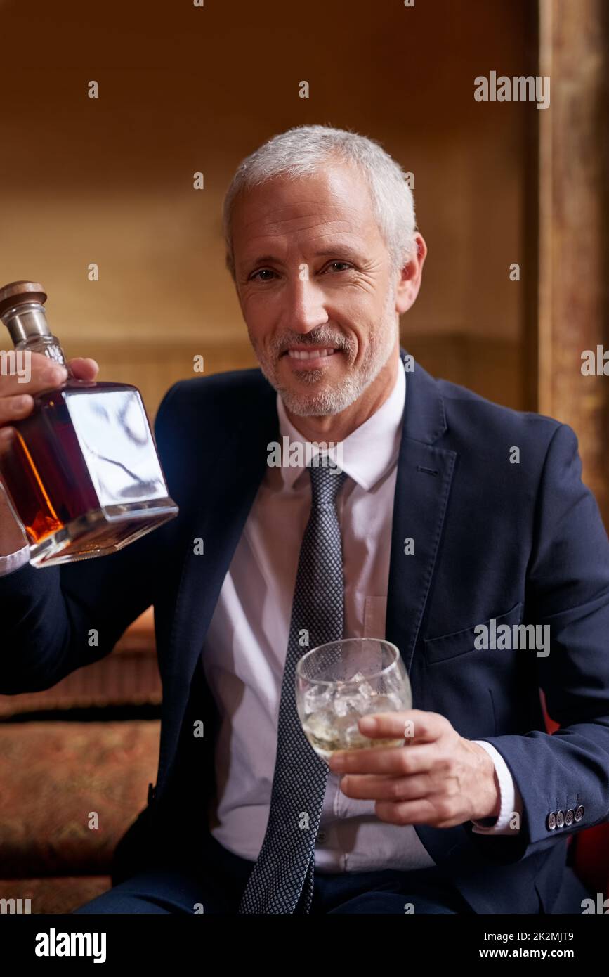 Es gibt nichts besseres als einen feinen Whiskey. Porträt eines gut gekleideten, reifen Mannes, der nach der Arbeit eine Glas- und Whiskey-Flasche in einer Bar hält. Stockfoto