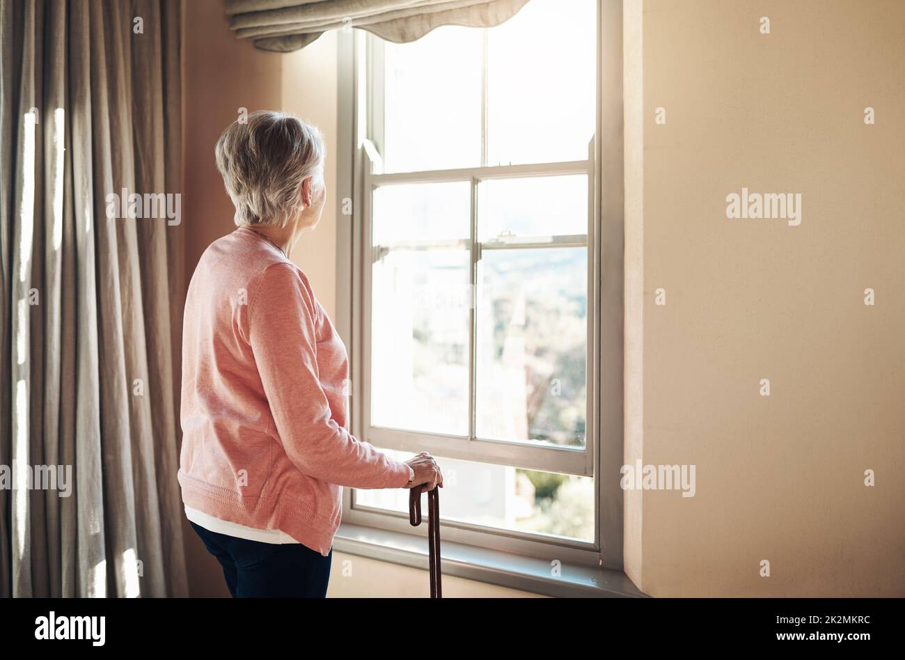 Wann wurden die Tage so grau. Aufnahme einer älteren Frau mit einem Stock, der zu Hause nachdenklich aus dem Fenster schaute. Stockfoto