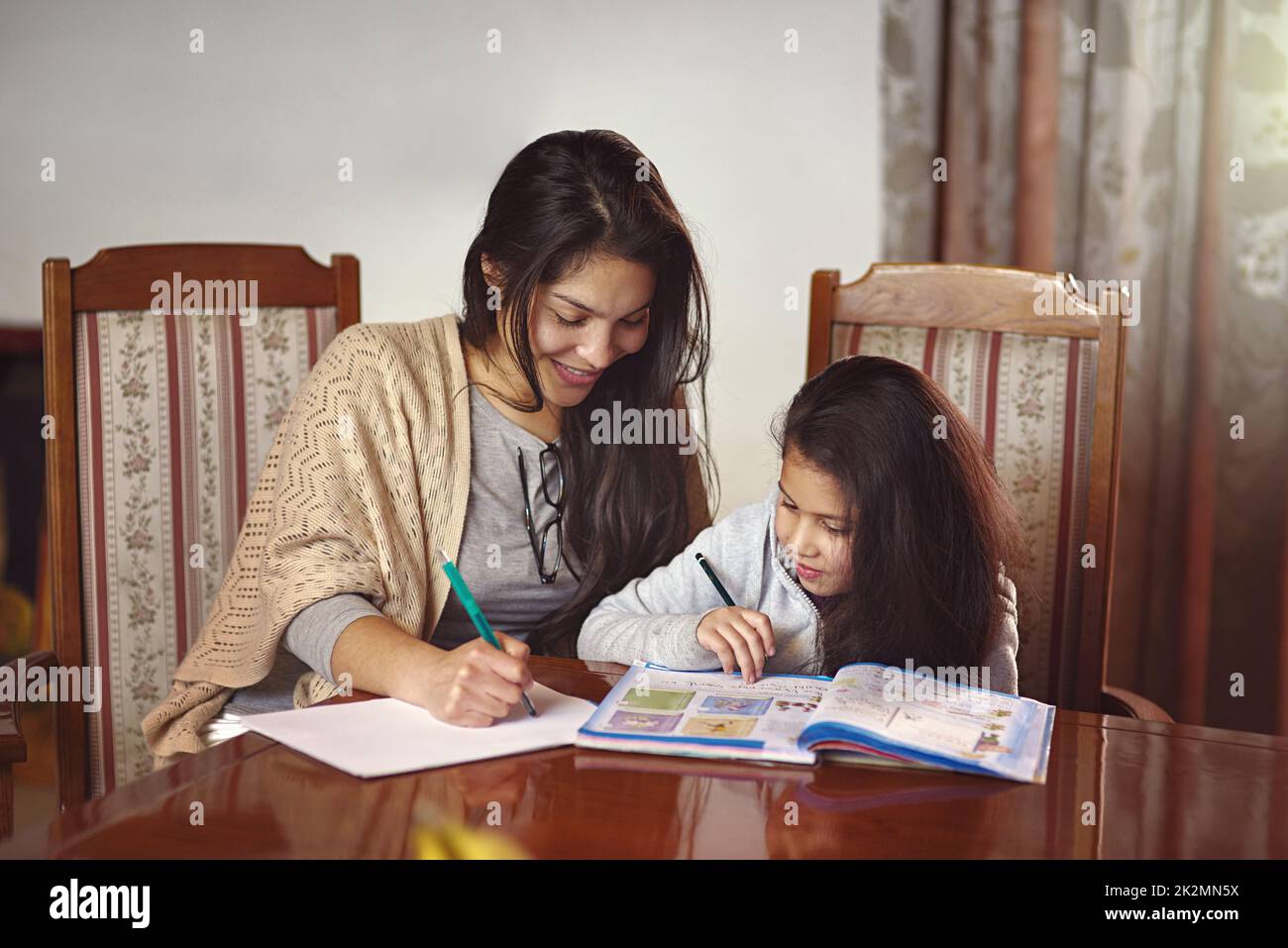Schaut, wie Mama es macht. Aufnahme einer Mutter, die ihrer Tochter zusieht, wie sie ihre Hausaufgaben zu Hause macht. Stockfoto