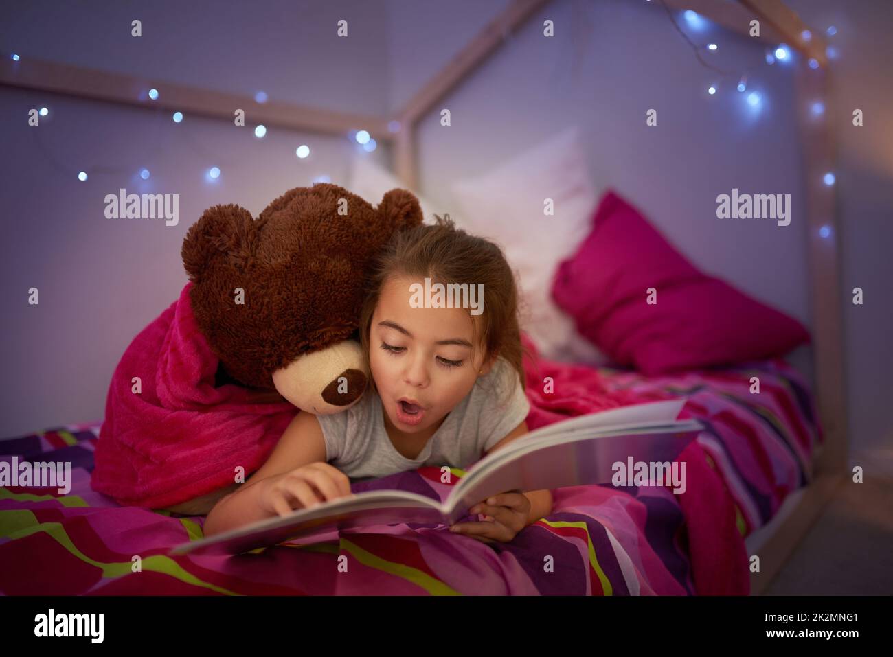 Es ist eine Geschichte voller wunderbarer Überraschungen. Eine kleine Aufnahme eines Mädchens, das mit ihrem Teddybär ein Buch im Bett liest. Stockfoto