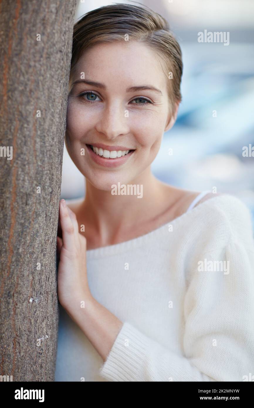 Es gibt so viele schöne Gründe, glücklich zu sein. Porträt einer lächelnden jungen Frau, die sich draußen an einen Baum lehnt. Stockfoto