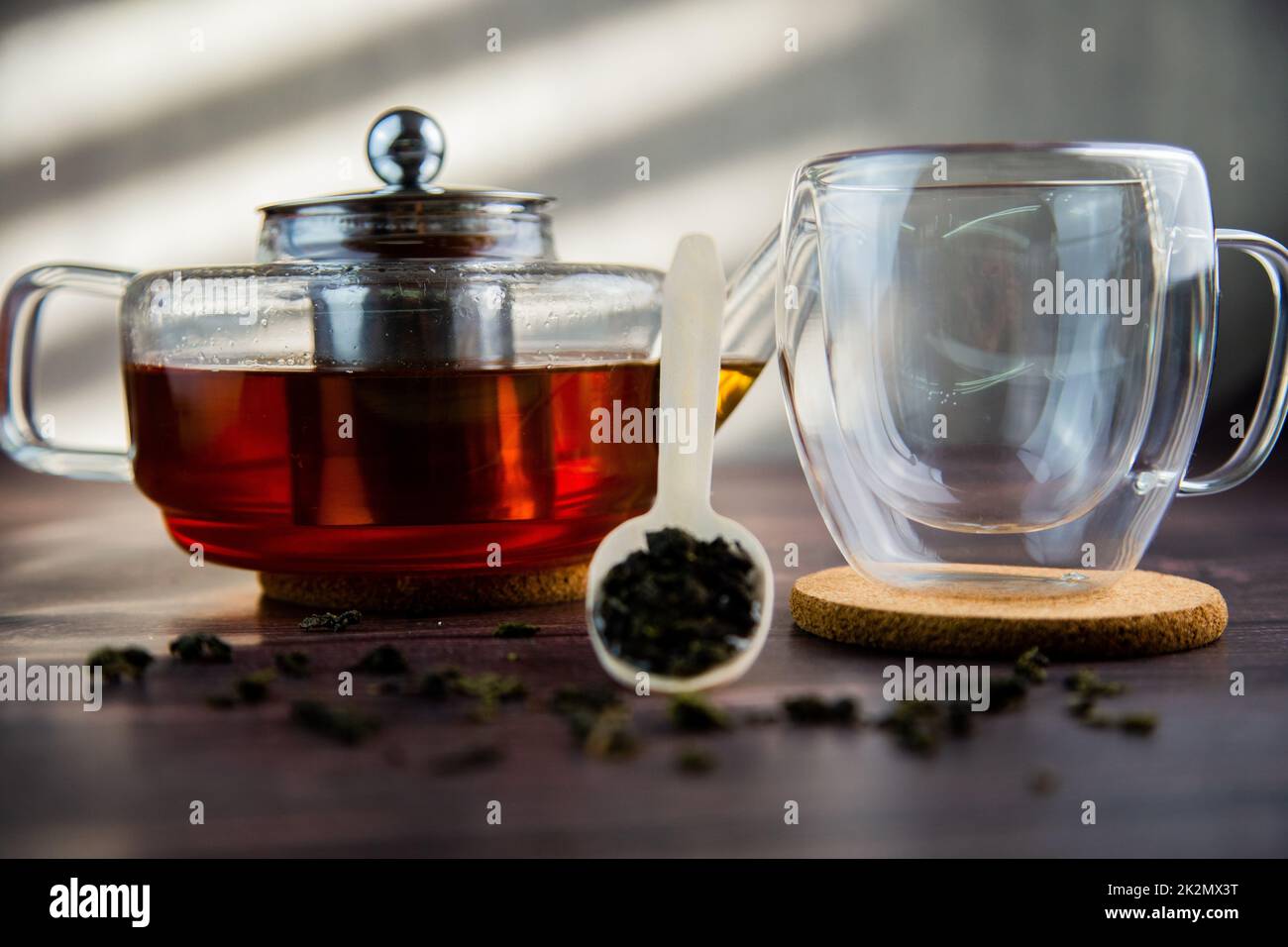 Ein Holzlöffel mit Tee liegt vor einem dunklen Holzhintergrund, ein Becher und eine Teekanne sind von hinten sichtbar, ein Foto in dunklen Farben. Stockfoto