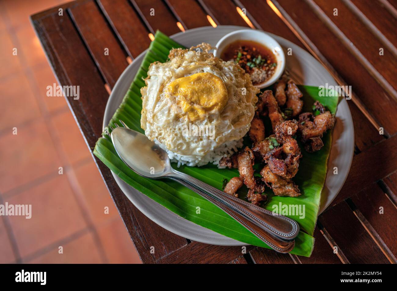 Knuspriger Schweinebauch mit Spiegelei, gedünstetem Reis und süßer würziger Sauce in Keramikschale auf Holztisch serviert Stockfoto