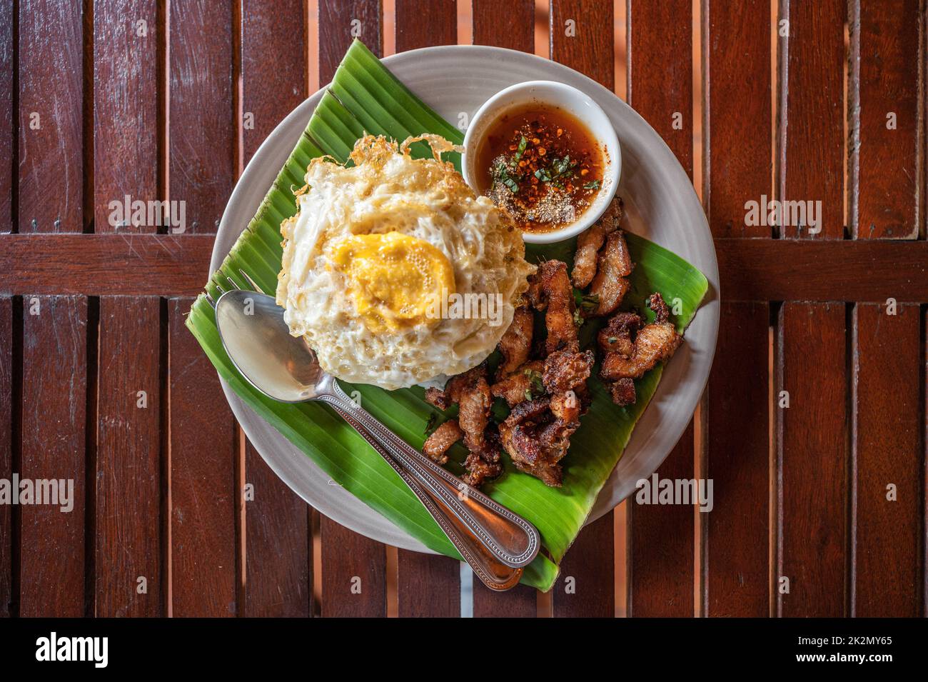 Knuspriger Schweinebauch mit Spiegelei, gedünstetem Reis und süßer würziger Sauce in Keramikschale auf Holztisch serviert Stockfoto