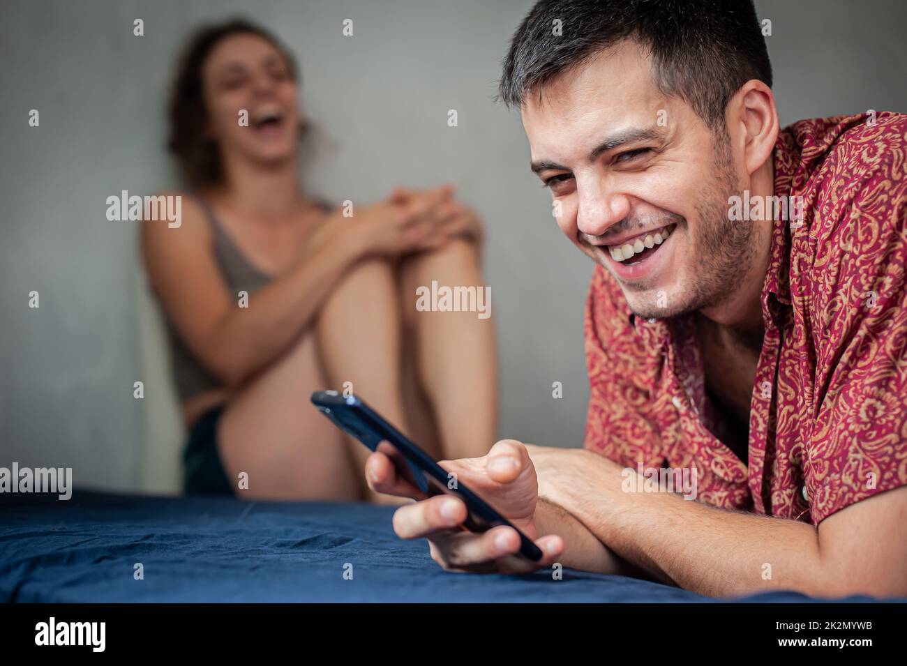 Ein junges, fröhliches Paar liegt und sitzt auf dem Bett im Schlafzimmer und nutzt ein Smartphone zur Kommunikation und Information Stockfoto