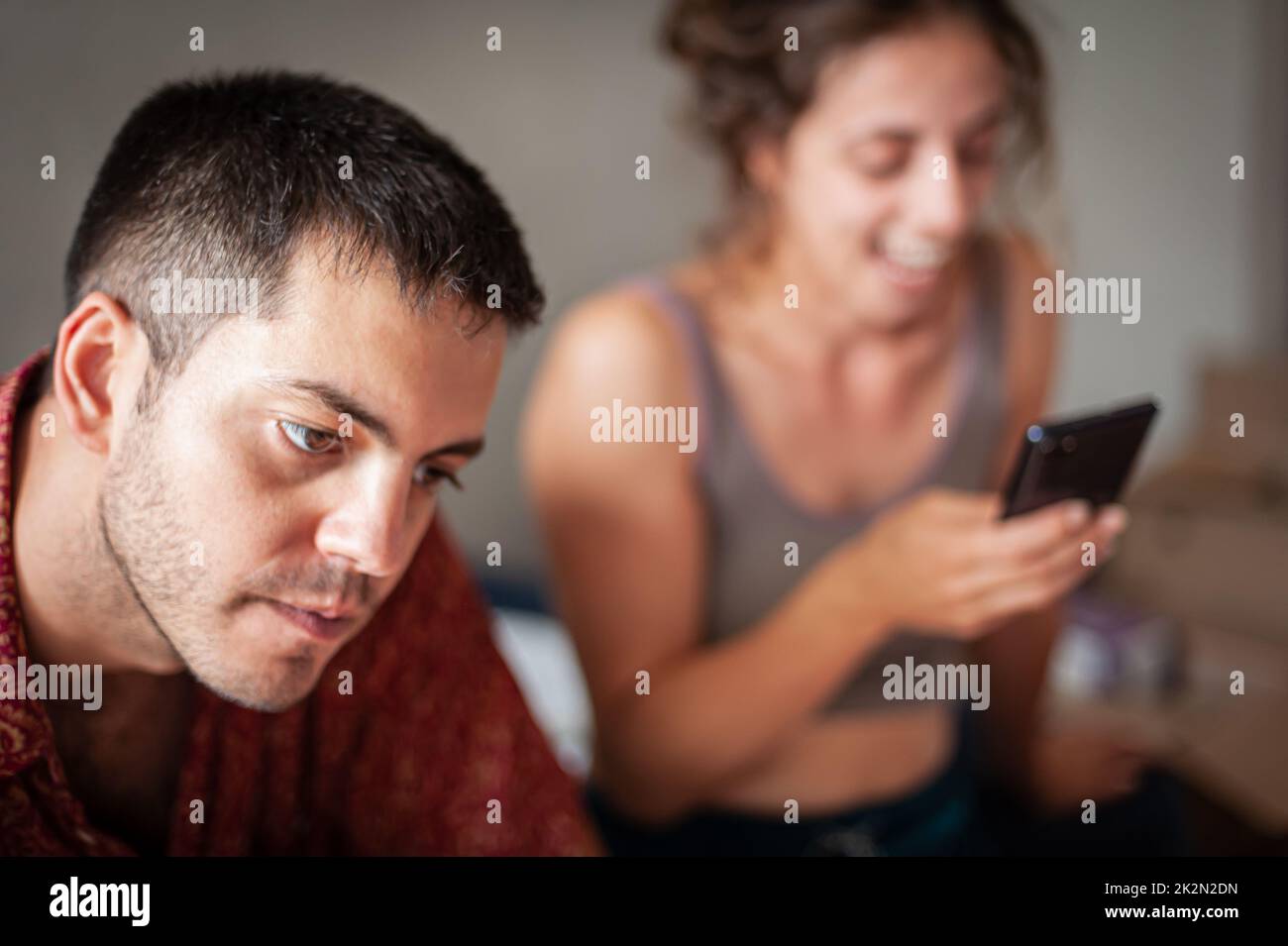 Ein junges, fröhliches Paar liegt und sitzt auf dem Bett im Schlafzimmer und nutzt ein Smartphone zur Kommunikation und Information Stockfoto