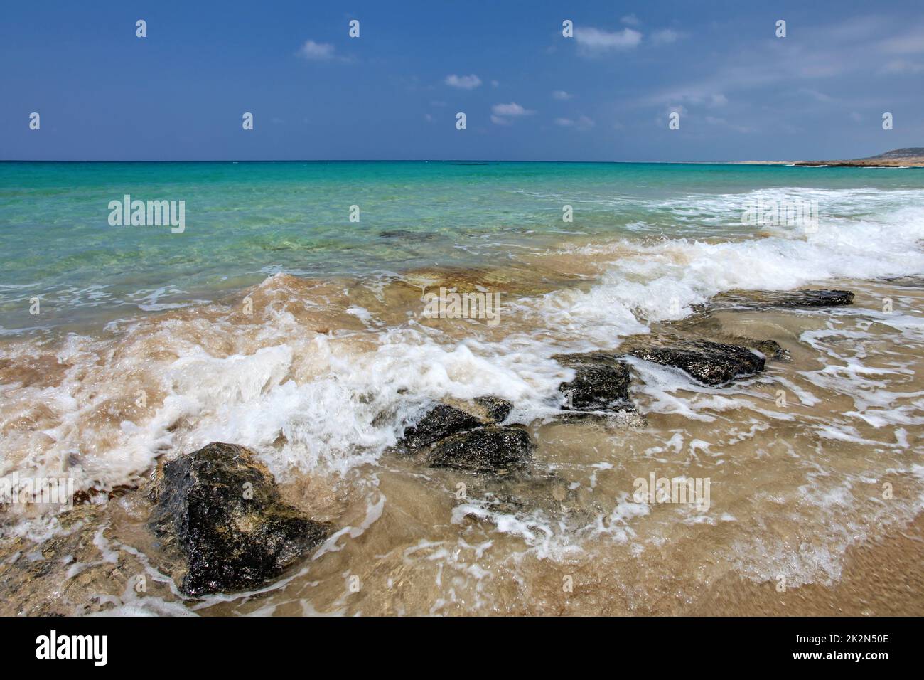 Kleine Wellen brechen am schwarzen Felsen am sandigen Strand, schöne blaue kristallklare Wasser im Hintergrund. Karpass, Nordzypern Stockfoto