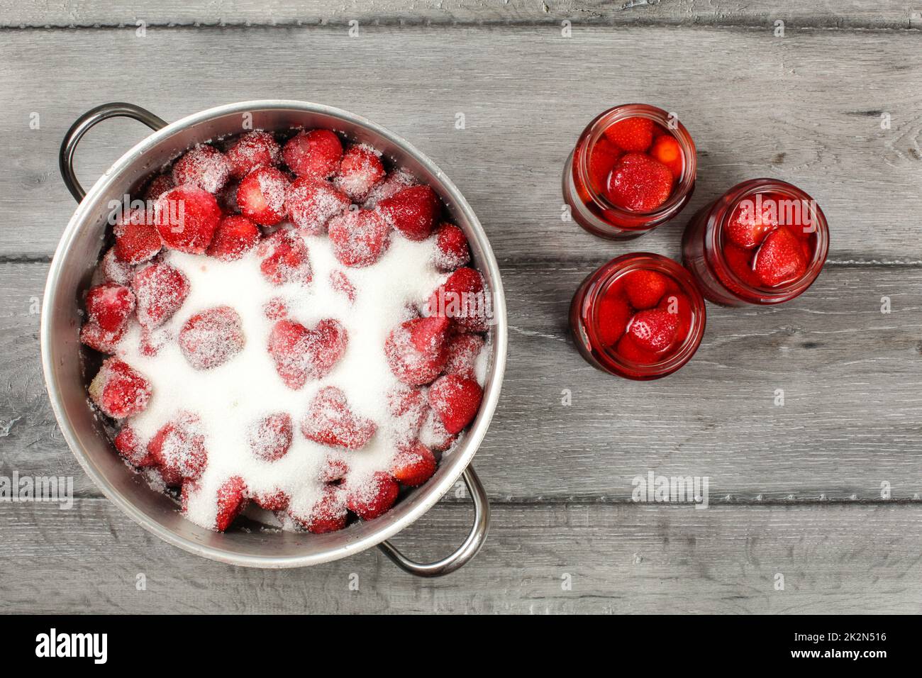 Blick auf den Tisch - großer Stahltopf mit Erdbeeren mit Kristallzucker, drei eingelegte Erdbeerglasflaschen daneben. Hausgemachte Kompottzubereitung. Stockfoto