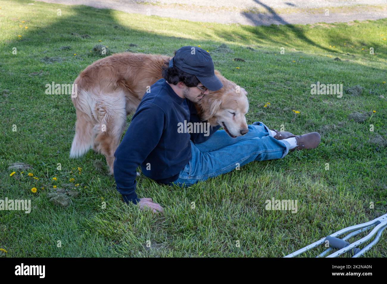 Der Golden Retriever-Hund kuschelt mit einem Mann, der mit Krücken auf dem Rasen sitzt Stockfoto