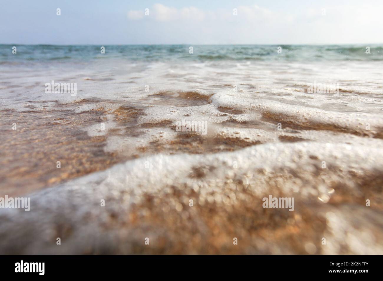 Niedriger Winkel, Kamera auf dem Boden, Objektiv mit Wassertropfen bedeckt, um Nässe hervorzuheben - Nahaufnahme von flachen Meereswellen, die den Sand am Strand waschen. Abstrakter Meereshintergrund. Stockfoto
