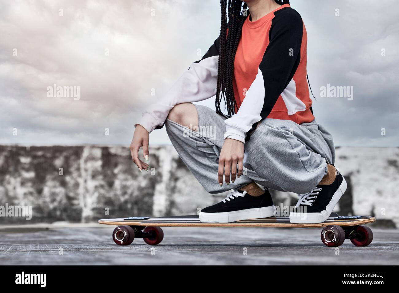 Sie sehen mich Rollen Eine kurzgeschnittene Aufnahme einer unerkennbaren jungen Skaterin, die vor stürmischer Kulisse auf ihrem Longboard auf einem Dach hockte. Stockfoto