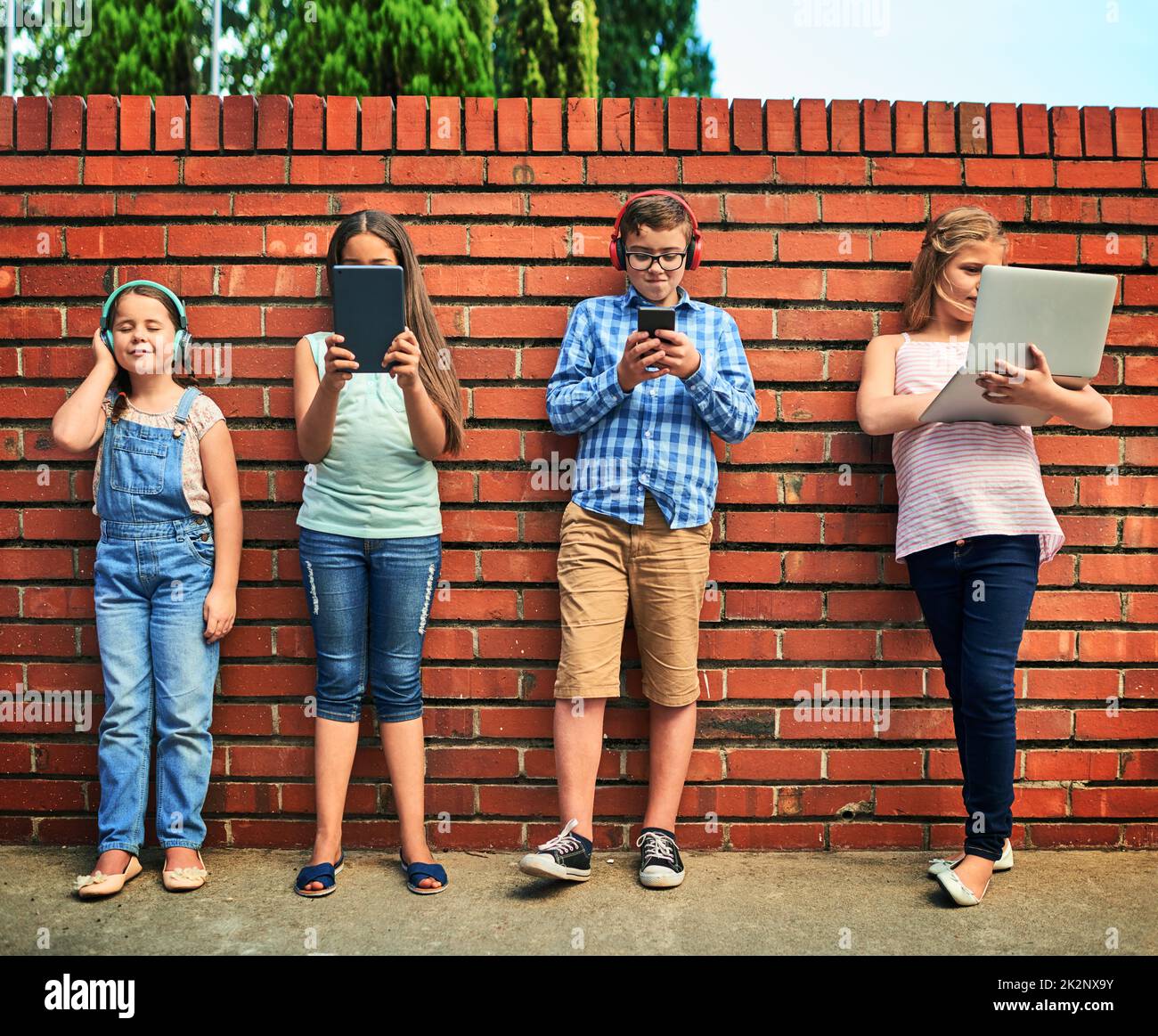 Kindheit im digitalen Zeitalter. Aufnahme einer Gruppe von kleinen Kindern, die digitale Geräte gegen eine Ziegelwand verwendeten. Stockfoto