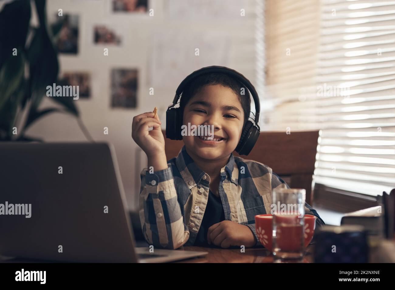 Die Pausenzeit ist die Snack-Zeit. Aufnahme eines entzückenden kleinen Jungen, der einen Laptop und Kopfhörer benutzt, während er zu Hause einen Schulauftrag abschließt. Stockfoto