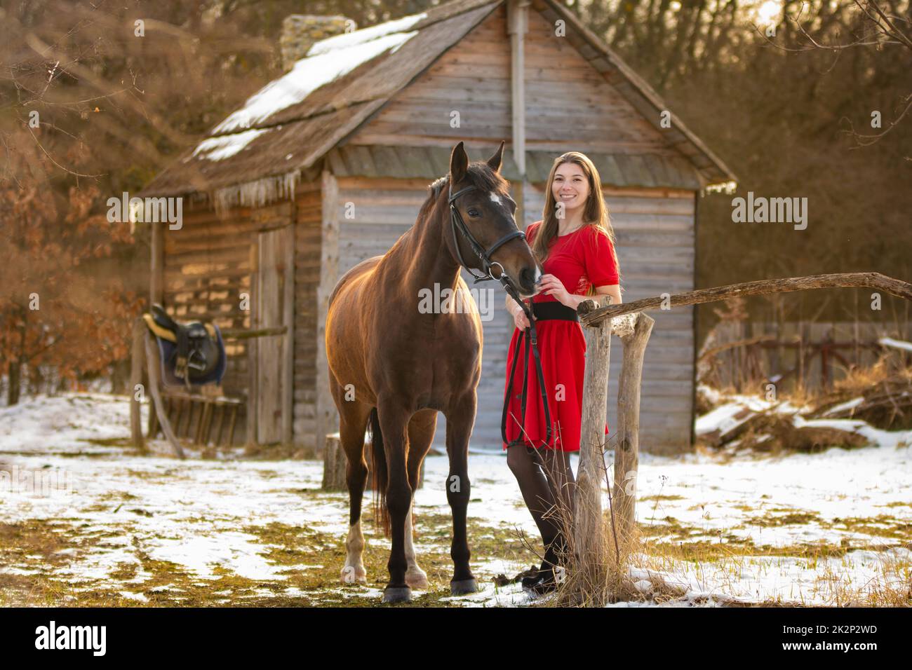 Ein schönes Mädchen in einem roten Kleid geht im Winter in den Strahlen der untergehenden Sonne durch einen alten Bauernhof Stockfoto