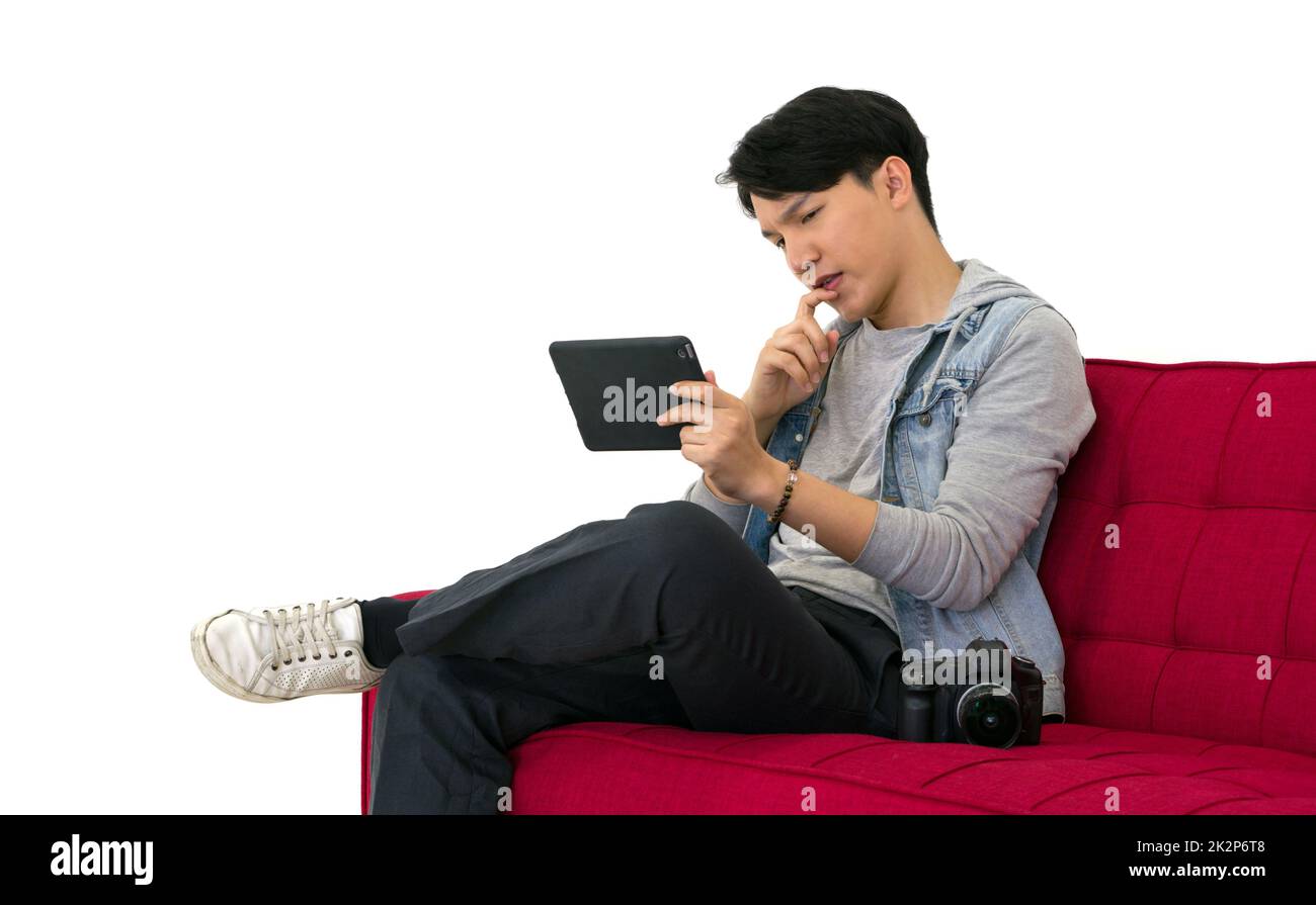 Der junge asiatische Fotograf sieht sich Bilder an, die auf dem Tablet-Computer-Bildschirm aufgenommen wurden, während er auf einem roten Sofa sitzt. Arbeitsatmosphäre im Fotostudio Stockfoto