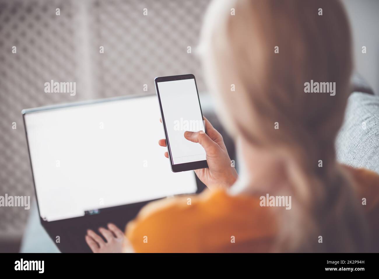 Frau mit Smartphone in den Händen sitzt drinnen und tippt auf dem Laptop Stockfoto
