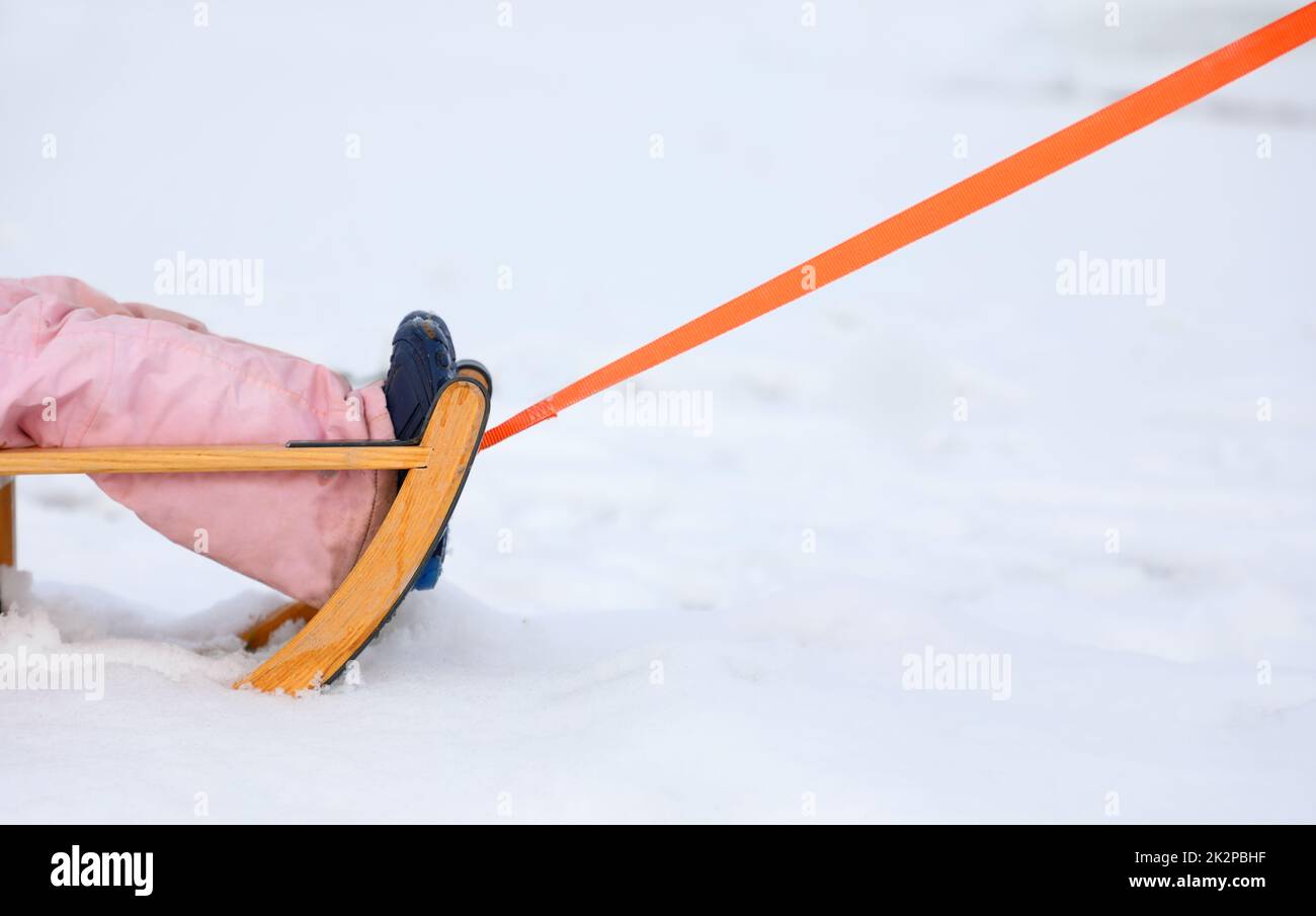 Die Spitze eines Schneelittens mit den Beinen eines Kindes, das auf einem verschneiten Hintergrund gezogen wird. Stockfoto