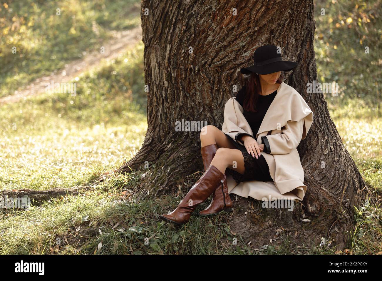 Frau in einem beigefarbenen Fell und schwarzen Hut sitzen unter Baum Stockfoto