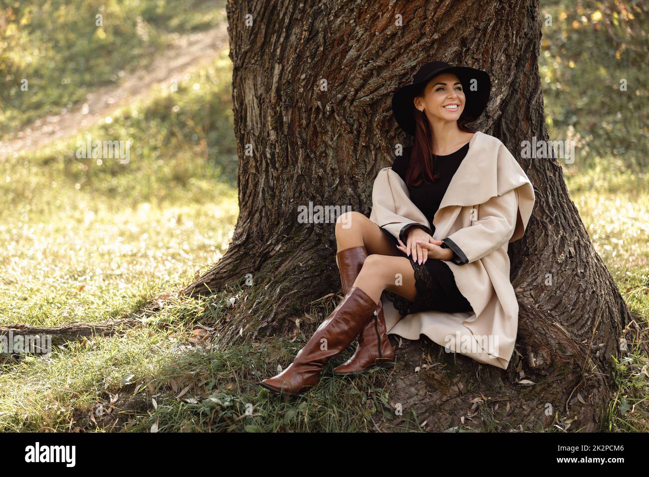 Frau in einem beigefarbenen Fell und schwarzen Hut sitzen unter Baum Stockfoto