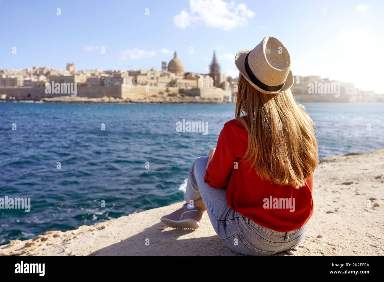 Reisekonzept mit unabhängigen Personen, die die Outdoor-Aktivitäten und den Lebensstil des Wanderessens genießen. Eine junge Frau sitzt am Meeresrand und sieht die atemberaubende Landschaft von Valletta, Malta, aus. Stockfoto