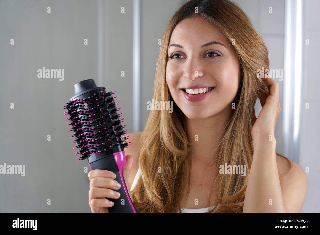 Erfreut Mädchen hält runde Bürste Haartrockner Haar in ihrem Badezimmer zu Hause Stil. Junge Frau zeigt einen einstufigen Haartrockner und Volumenizer im Salon. Stockfoto