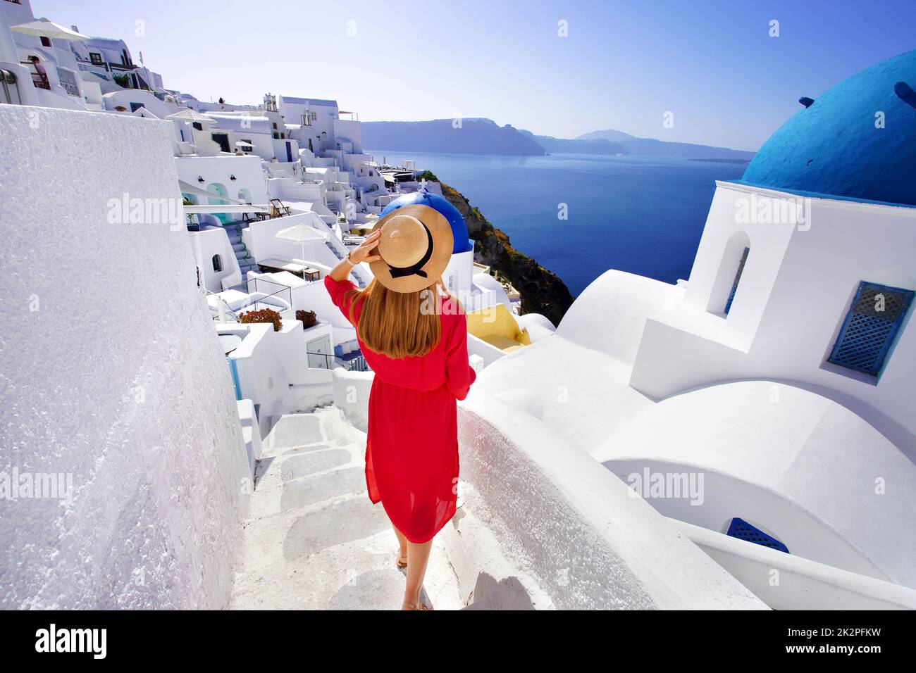 Schönes Mädchen in rotem Kleid und Hut kommt die Treppe hinunter und erkundet das Dorf Oia mit spektakulärem Blick auf die Caldera, Santorini, Griechenland Stockfoto