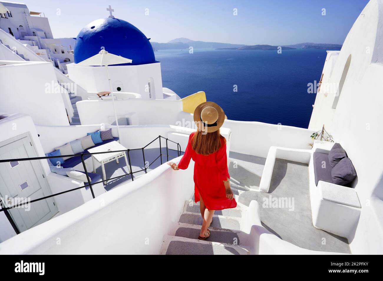 Schönes Mädchen in rotem Kleid und Hut kommt die Treppe hinunter zu ihrem Resort und genießt einen spektakulären Blick auf die Caldera im Dorf Oia, Santorini, Griechenland Stockfoto