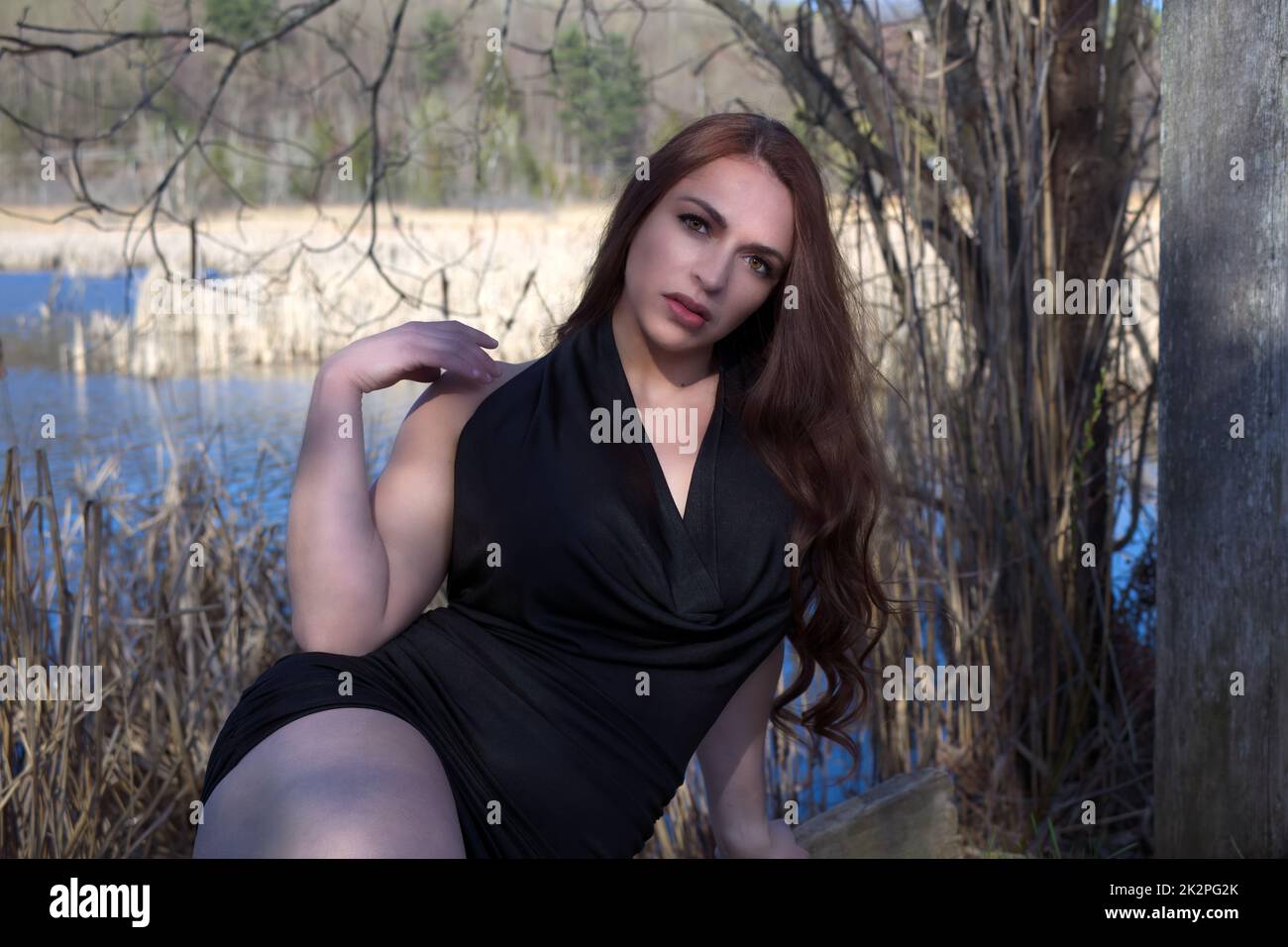 Junge Frau in schwarzem Kleid, die im Wald neben einem Wasserteich in der Sonne sitzt Stockfoto