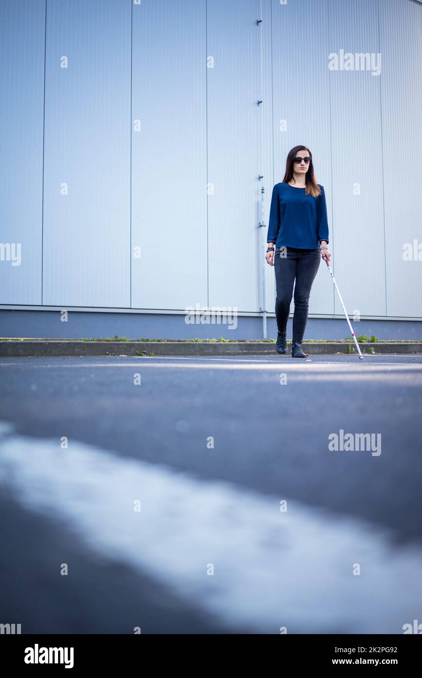 Junge Frau mit Sehbehinderung geht auf den Straßen der Stadt und nutzt ihren weißen Stock, um besser durch den städtischen Raum zu navigieren und sicher an ihr Ziel zu gelangen Stockfoto