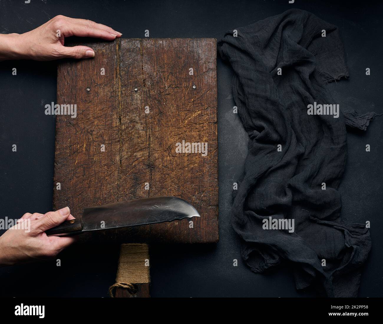 Leere braune rechteckige Holzschneidplatte, Frau hält großes Messer über dem schwarzen Tisch, Draufsicht Stockfoto