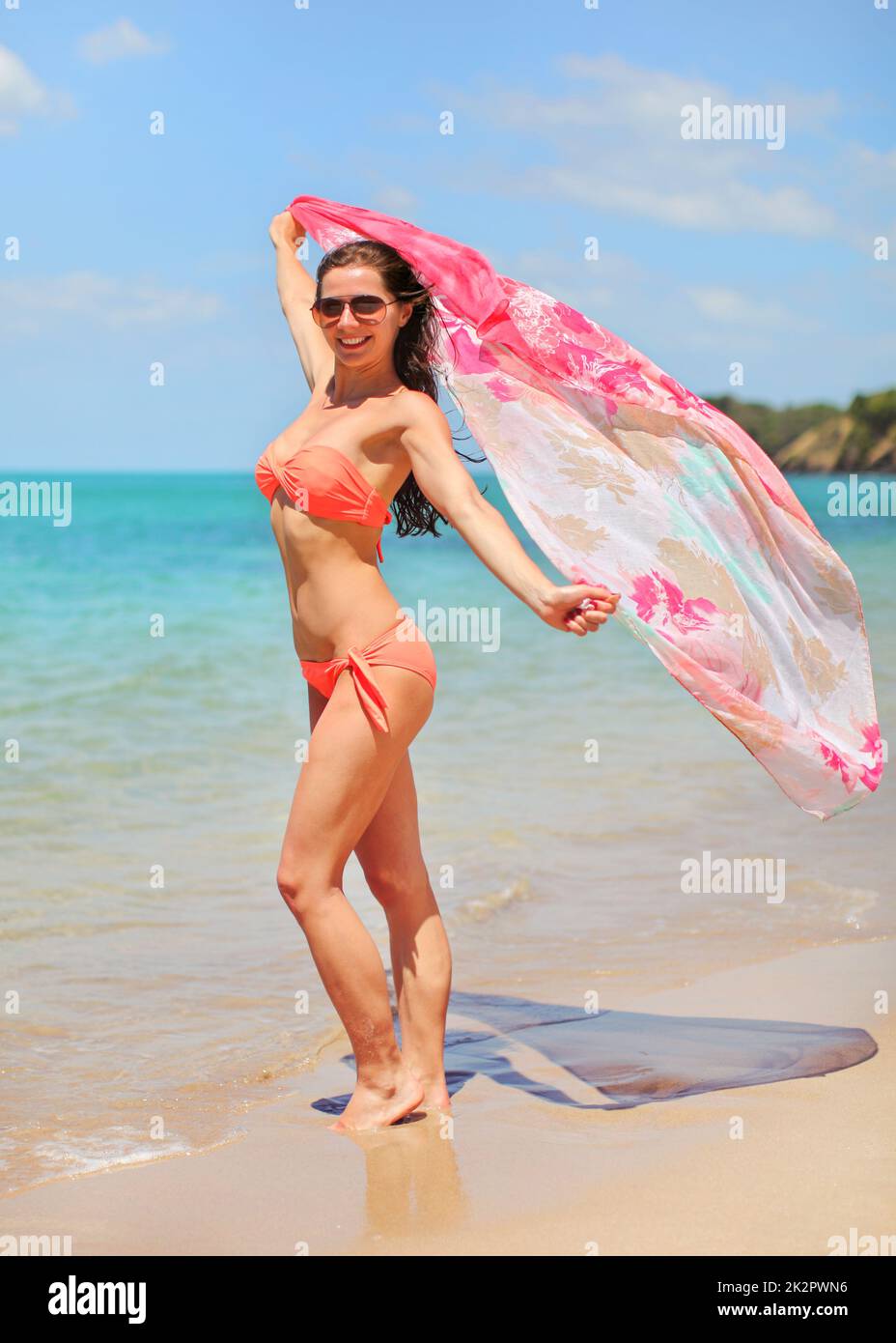 Eine junge, sportliche schlanke Frau in orangefarbenem Bikini und Sonnenbrille steht am Strand und hält hinter ihr einen Schal, der im Wind winkt, azurblauer Meereshintergrund. Stockfoto