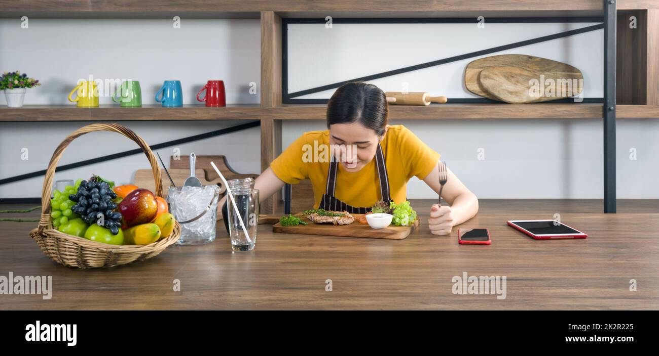 Die Hausfrau in einer Schürze gekleidet, bereitete eine Gabel und ein Messer, bereit, ein leckeres Steak zu essen. Morgenstimmung in einer modernen Küche Stockfoto