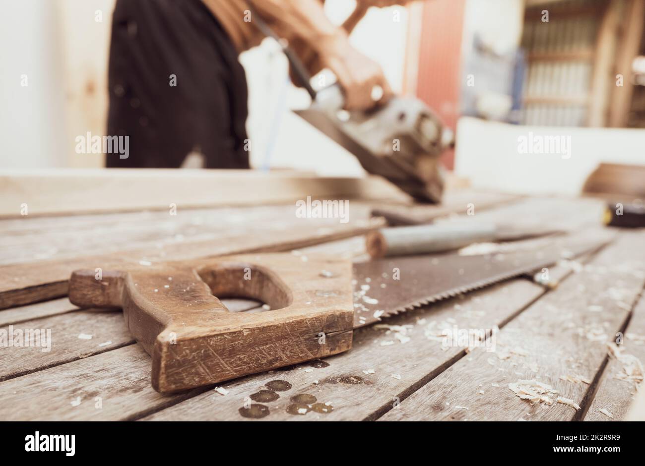 Handsäge mit Holzgriff auf verschwommenem Tischler, der mit elektrischer Hobelmaschine arbeitet. Zimmermannswerkzeuge. Handsäge und Sägemehl auf Holztisch in der Außenwerkstatt. Ein Handwerker, der Holzarbeiten macht. Handwerkkonzept. Stockfoto