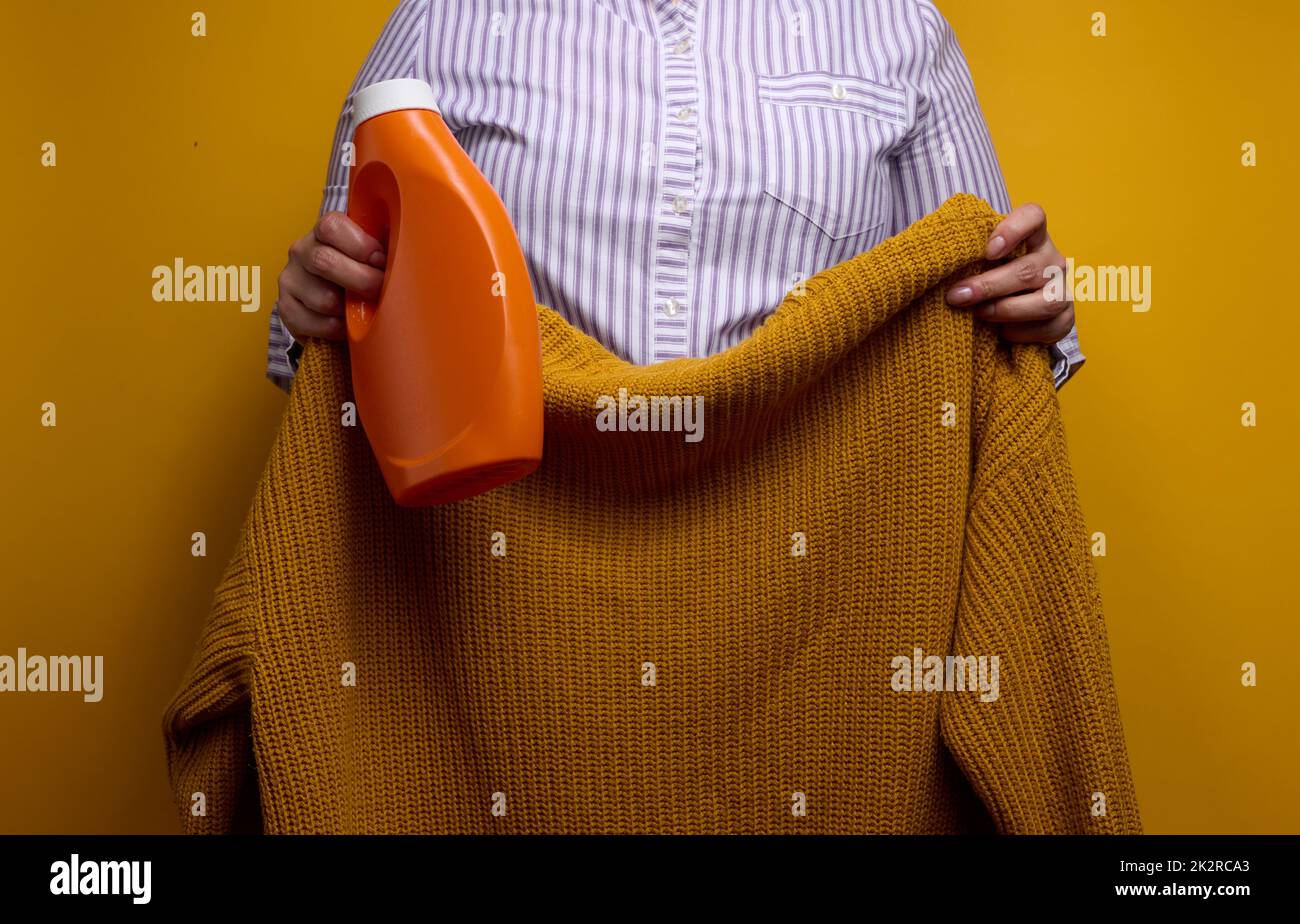 Eine Frau in einem weißen gestreiften Hemd hält eine orangefarbene Plastikflasche mit flüssigem Waschgel und einen Strickpullover. Wäsche- und Haushaltsarbeiten, saubere Wäsche Stockfoto