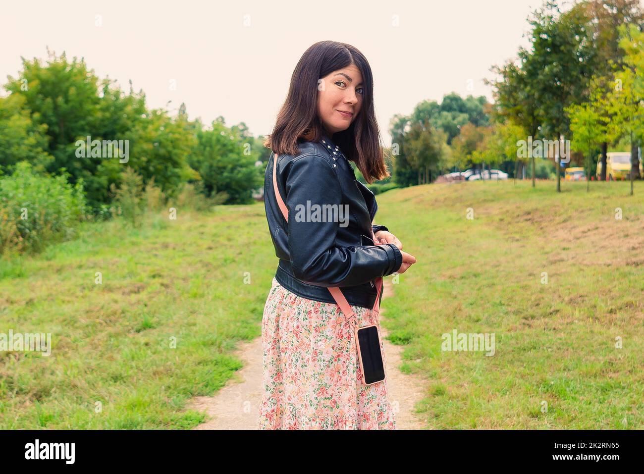 Ein schönes lächelndes Mädchen mit einem Telefon am Gürtel blickt zurück, während es mitten in einem grünen Park steht Stockfoto