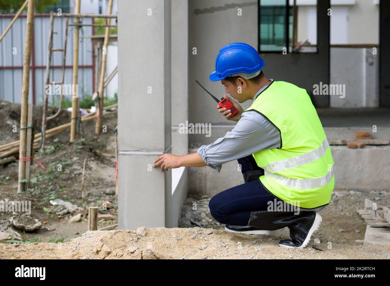 à¹à¹Young asiatischer Mann mit Schutzhelm und Sicherheitsweste, der die Hausstruktur überprüft, während er den Walkie-Talkie hält. Arbeitsumfeld von Ingenieuren auf der Baustelle von Wohnungsbauprojekten Stockfoto