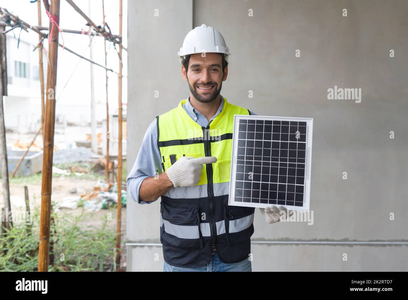 Der junge Ingenieur in Schutzweste und Handschuh zeigt mit dem Finger auf die Solarzellenplatte und stellt mit einem Lächeln eine Energiequelle dar, um Gleichstrom zu erzeugen. Stockfoto